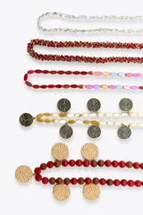 Pack de cinco collares superpuestos con cadenas de diferentes grosores. Incluye collares con cuentas rojas, marrones y amarillas, cuentas de cristal transparentes/rojas, y dijes metálicos circulares con textura martillada dorada y otros con diseño de espiral plateado/negro.