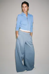 Pantalón wide leg de tiro medio confeccionado en hilatura de algodón, color beige, con trabillas en la cintura, bolsillos laterales ocultos y detalle de pliegues en el delantero. Cierre frontal con cremallera, botón y ganchos metálicos.
