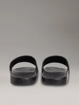 Ojotas tipo slide de color negro con tira superior de piel sintética que presenta un patrón grabado del logo de Calvin Klein. Poseen plantilla ergonómica moldeada y suela de EVA ligera.