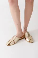 Zueco de cuero color dorado con diseño de tiras anchas entrelazadas y punta cuadrada. Calzado plano tipo mule sin talón, diseñado para la temporada de verano.