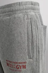 Pantalón jogger gris de corte regular fit, confeccionado en tejido de algodón con interior afelpado. Cuenta con cintura elástica ajustable con cordón, bolsillos laterales de vivo y traseros de plastrón. Presenta un texto estampado en color bordó en la pernera y bajo acabado en puño con elástico. Incluye un parche con logo combinado a contraste en la cintura.
