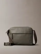 Bolso bandolera Calvin Klein de cuero sintético texturizado color verde oliva, con correa ajustable de tela marrón oscuro que lleva el logo de la marca repetido. Presenta un bolsillo frontal horizontal y el logo CK grabado en la parte inferior derecha.