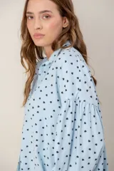 Camisa de corte oversize y diseño holgado, color celeste con estampado de pequeños corazones negros. Presenta cuello clásico, cierre frontal con botones y mangas largas abullonadas con puños ajustados.