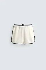 Short deportivo de tejido de punto con estructura abierta, color blanco con vivos a contraste en azul marino. Presenta cintura elástica, bolsillos frontales, aberturas laterales en el bajo y etiquetas conmemorativas de la Copa Mundial de la FIFA Francia 98.