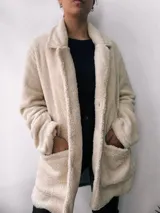 Saco de abrigo color crudo, de corte recto y largo a la cadera, confeccionado en tejido tipo teddy con cuello solapa y cierre frontal con botones.