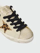 Championes Golden Goose modelo Super Star, con diseño de efecto desgastado. Presentan cuerpo en cuero color crema, puntera y lengüeta con acabado de glitter dorado, estrella lateral con estampado animal print y cordones negros.