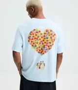 Remera boxy de algodón color celeste, con cuello redondo y mangas cortas. Presenta un estampado frontal con lettering y un estampado trasero grande con un corazón formado por círculos coloridos y una pequeña ilustración de una casa debajo, inspirado en la película "Up: Altas Aventuras".