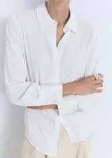 Camisa blanca de tejido fluido con cuello camisero, manga larga con puños abotonados y cierre frontal de botones ocultos.