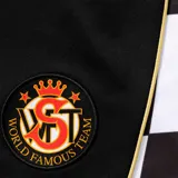 Short deportivo negro con paneles laterales a cuadros blancos y negros. Presenta detalles en color dorado, el logo de Supreme en la parte inferior de los laterales y un escudo circular bordado en la pierna derecha.