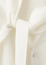 Blusa de modal y poliéster color blanco marfil, con diseño recto, escote en V, mangas largas y detalle de fular incorporado en el cuello. Cierre frontal con botones.