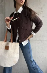 Cardigan de punto color chocolate, con diseño de trenzas y pompones decorativos. Presenta cuello redondo, cierre frontal con botones metálicos dorados y mangas largas.
