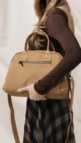 Cartera de mano color beige, confeccionada en tela impermeable, con doble asa corta y correa larga ajustable y desmontable. Presenta cierre principal, bolsillo frontal con cierre y detalle de colgante decorativo.