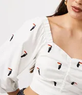 Blusa cropped de lino blanca con manga tres cuartos, hombro asimétrico, escote corazón, lastex en la espalda y bordados de tucanes.
