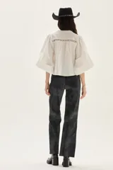 Blusa de algodón color blanco con mangas abullonadas, cuello mao y cierre frontal con botones. Presenta detalles de encaje tipo vainilla en el frente y en la parte superior del pecho.