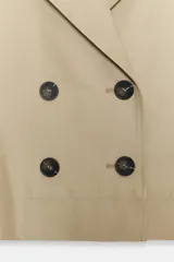 Trench corto de color beige, con cuello de solapa, cierre cruzado con botones y mangas cortas con vuelta y trabilla con botón.