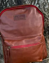 Mochila de cuero negro, con forro interior de lino y herrajes de bronce. Presenta un bolsillo frontal con cierre y un parche de cuero marrón con logo grabado.