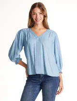 Blusa de lyocell color celeste, con escote en V y detalle de pespuntes verticales en el centro. Presenta mangas tres cuartos abullonadas con puños elásticos y volados, y frunces en los hombros.