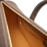 Cartera tipo baúl compacta, color marrón, con material exterior de símil gamuza combinado con PU liso. Cuenta con doble asa corta rígida, correa larga regulable y desmontable, y herrajes metálicos dorados. El compartimento principal tiene cierre superior y un bolsillo interno con cierre. Incluye detalle frontal con logo XL metalizado.