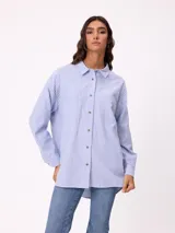 Camisa de corte oversize con diseño a rayas verticales en tonos azul y blanco. Presenta cuello clásico, cierre frontal con botones y un bolsillo en el pecho con detalle de etiqueta.