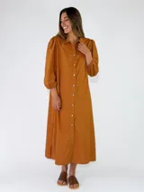 Vestido largo estilo camisero color tostado, con cierre frontal de botones, cuello camisero y mangas largas abullonadas con detalles bordados.