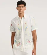 Camisa blanca de lino y algodón, de corte comfort, cuello inglés y abotonado frontal. Presenta manga corta y un bordado floral multicolor con motivos de aves y flores.
