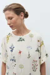 Blusa de manga corta, color crudo, con estampado floral en tonos azules, amarillos y rojos. Presenta cuello redondo y volados en los puños.