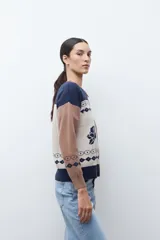 Chaqueta de tejido punto con cierre frontal, cuello redondo y manga larga. Presenta un diseño jacquard en tres colores: azul marino, beige y marrón camel. El cuerpo principal es beige con motivos de rosas azules y patrones geométricos en azul marino. Los hombros y mangas son de color marrón camel con patrones geométricos en azul marino. El cuello y el elástico inferior son de color azul marino.