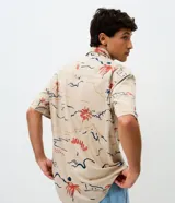 Camisa de corte relajado con cuello solapa y manga corta, confeccionada en algodón con estampa abstracta de palmeras y paisajes en tonos tierra, azul y rojo.