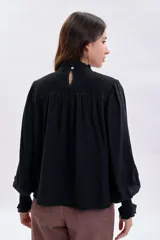 Blusa negra de corte holgado con detalle de punto smock en el pecho y cuello alto con volado. Presenta mangas largas abullonadas con puños elásticos fruncidos.