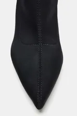 Botín de tejido elástico color negro con acabado en punta y tacón fino alto. Presenta una tira decorativa con hebilla metálica en la caña y cierre mediante cremallera lateral.