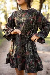 Vestido corto de lino con estampado floral en tonos tierra y verdes. Presenta cuello redondo, mangas largas amplias con puños terminados en volados y falda con volado en el ruedo. Incluye un lazo desmontable para ajustar la cintura.