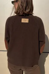 Saco de punto color beige, con cierre frontal de botones marrones y detalle de costuras decorativas oscuras en los hombros. Presenta un diseño clásico con cuello redondo.
