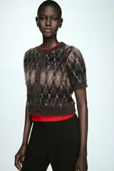 Chaqueta de punto de manga corta y cuello redondo, con cierre frontal de botones y diseño de rombos en tonos marrones, negros y blancos.
