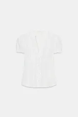 Camisa de popelín con cuello redondo y escote en pico, manga corta abullonada con elástico y cierre frontal con botones. Presenta detalles de volantes a lo largo de la abertura frontal.