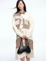 Vestido camisero de corte midi con diseño patchwork que combina paneles en tonos crudo y marrón con estampados florales sutiles. Presenta cierre frontal con botones, cuello camisero y mangas largas con puños.