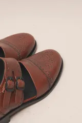 Zapato tipo monk strap con aberturas laterales, confeccionado en cuero vacuno metalizado color bronce. Presenta detalles de perforaciones estilo brogue en la puntera y el talón, y doble hebilla metálica en el empeine.