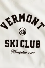 Buzo oversized color crema con cuello redondo acanalado. Presenta un estampado frontal en negro con el texto "VERMONT SKI CLUB Montpelier 1970" y un logo de montaña. Tiene hombros caídos y remates acanalados en puños y bajo.