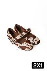 Chatas estilo Mary Jane confeccionadas en cuero con pelo, con estampado de manchas tipo vaca en tonos marrón y blanco. Presentan una tira ajustable sobre el empeine con hebilla metálica.