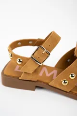 Sandalia tipo cangrejera de gamuza color camel, con diseño de tiras entrelazadas y tachas metálicas doradas. Cuenta con cierre de pulsera con hebilla ajustable y suela plana de goma.