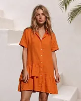 Vestido camisero corto de lino color blanco, con mangas cortas, cierre frontal de botones, cuello camisero y volados asimétricos en el ruedo.
