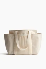 Cartera shopper grande confeccionada en tejido grueso de mezcla de yute color beige, con doble asa de hombro, compartimentos de malla en el frente y dorso, y bolsillos laterales abiertos.