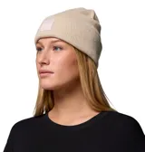Gorro tipo beanie de tejido grueso color beige, con diseño de punto acanalado y vuelta en el borde. Presenta un parche cuadrado frontal con el logo de la marca Columbia.