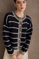 Cardigan de punto con diseño a rayas horizontales en azul marino y beige. Presenta cuello redondo, cierre frontal con botones grandes y mangas largas con puños acanalados en tono verde oliva. Incluye un pequeño bordado de una raqueta de tenis en el lateral.