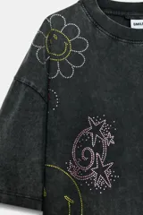 Camiseta oversize de algodón con efecto lavado, cuello redondo y manga corta. Presenta múltiples aplicaciones de brillos y strass con motivos coloridos de Smiley, incluyendo estrellas, corazones y caras sonrientes.