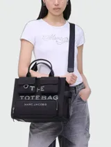 Cartera tote de lona color negro, modelo The Cargo Canvas Medium Tote Bag de Marc Jacobs. Presenta múltiples bolsillos frontales con solapa y cierre de cremallera, asas de mano superiores y una correa de hombro ajustable y extraíble. Cuenta con el nombre de la marca y la inscripción "THE TOTE BAG" estampados en el frente.