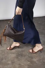 Cartera bandolera de cuero negro con flecos colgantes en el lateral, cierre superior y correa ajustable con argollas metálicas.