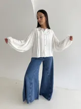 Blusa blanca con cuello mao y cierre frontal de botones de nácar. Presenta mangas largas abullonadas con puños elásticos y detalle de tablas verticales en el frente.