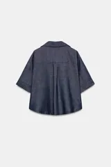 Camisa crop efecto denim color negro, con cuello solapa, escote en V, mangas cortas con trabilla y bolsillos frontales de parche. Cierre frontal con botones.