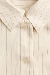 Camisa oversized de color beige con diseño de rayas finas, cuello clásico, cierre frontal de botones, bolsillo en el pecho y mangas largas con puños abotonados.