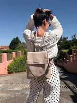 Mochila tipo cartera con tapa, confeccionada en símil cuero color suela. Cuenta con doble compartimento, división interna, cierre de seguridad y bolsillo trasero con cierre. Incluye una correa frontal decorativa con tachas metálicas.